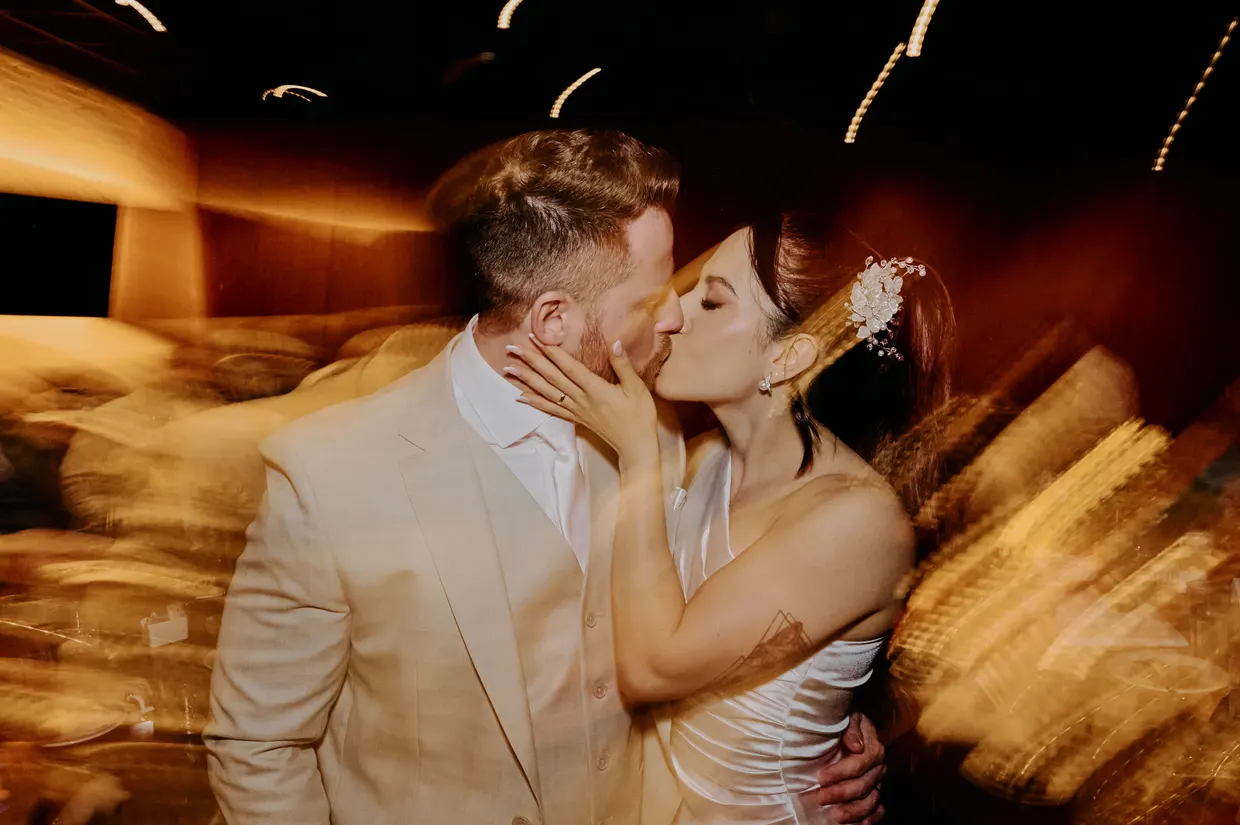 Casal se beija durante uma celebração, com luzes desfocadas ao fundo.