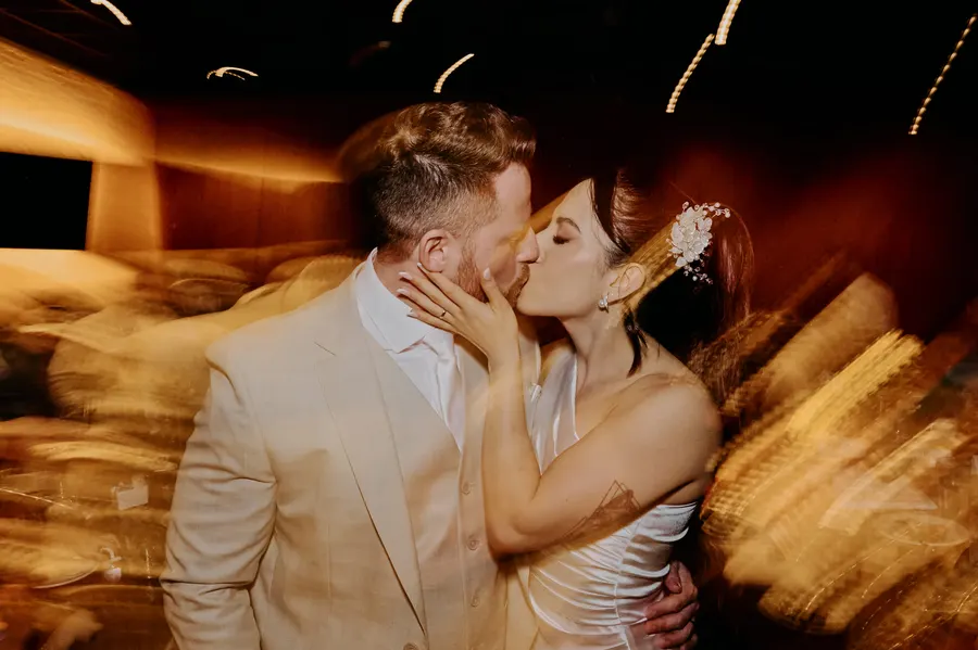 Casal se beija durante uma celebração, com luzes desfocadas ao fundo.