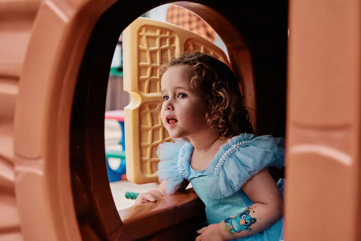 Criança de vestido azul olha pela janela de uma casinha de brinquedo.