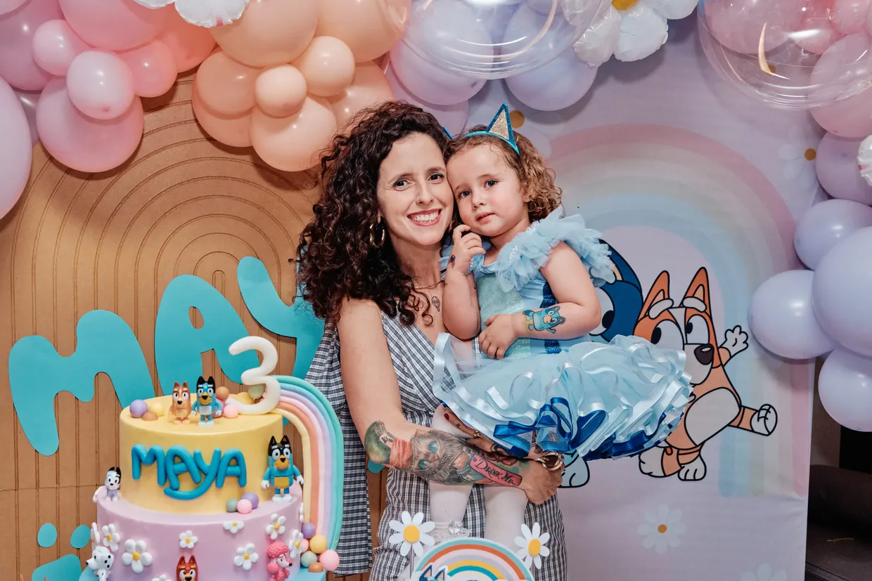 Mulher abraça uma criança diante de um painel de balões de aniversário.