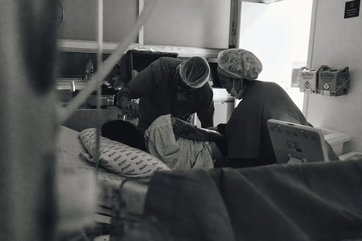 Fotografia em preto e branco de pessoas acompanhando um atendimento em quarto hospitalar.