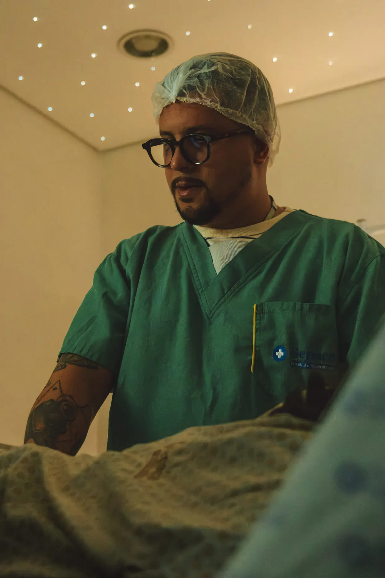 Homem de roupa hospitalar verde e touca acompanha uma cena de parto.