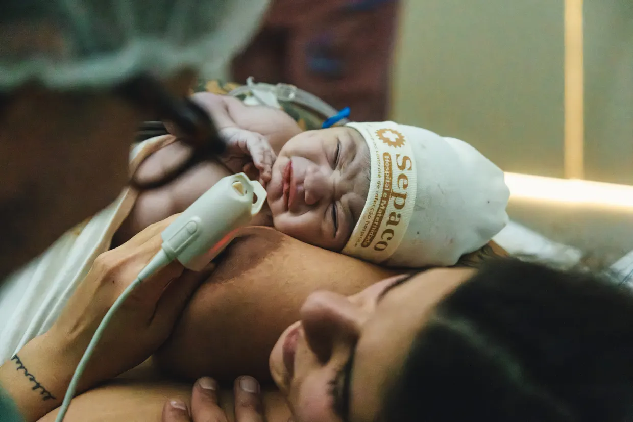 Mãe sorri com o recém-nascido de touca acomodado junto ao seu rosto.