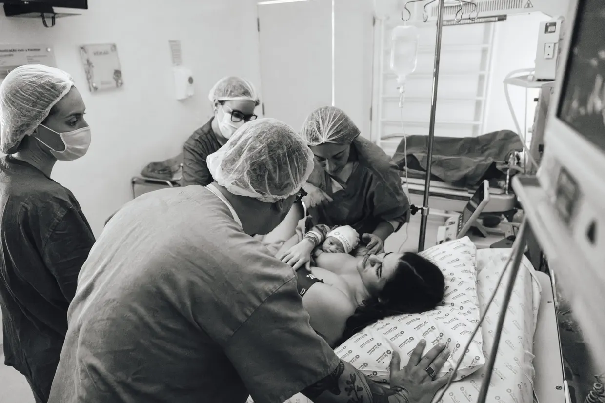 Fotografia em preto e branco de pessoas reunidas ao redor da mãe e do bebê no hospital.