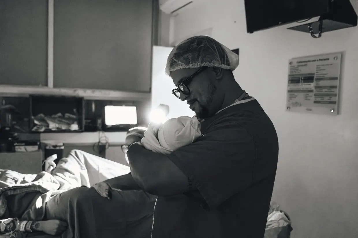 Pessoa de roupa hospitalar segura um bebê no colo, em fotografia em preto e branco.