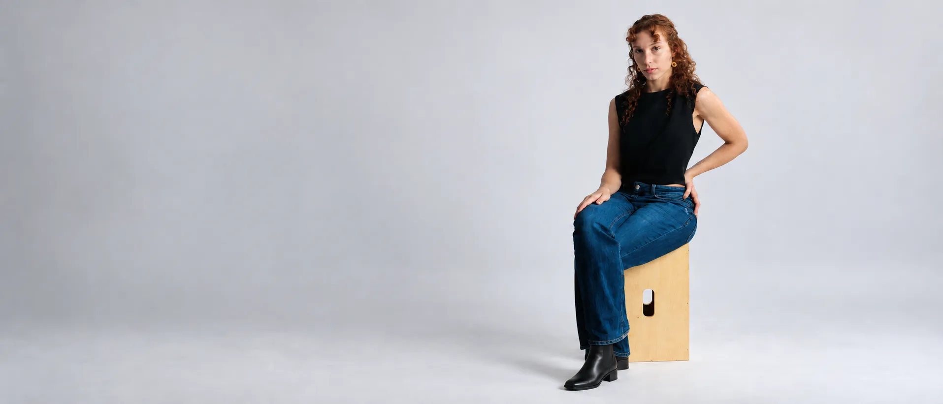 Retrato em estúdio: mulher sentada em um banco de madeira, com blusa preta, jeans e botas.