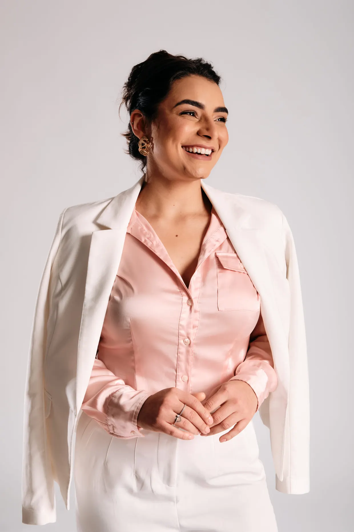 Mulher sorrindo, com camisa rosa e blazer claro apoiado nos ombros.