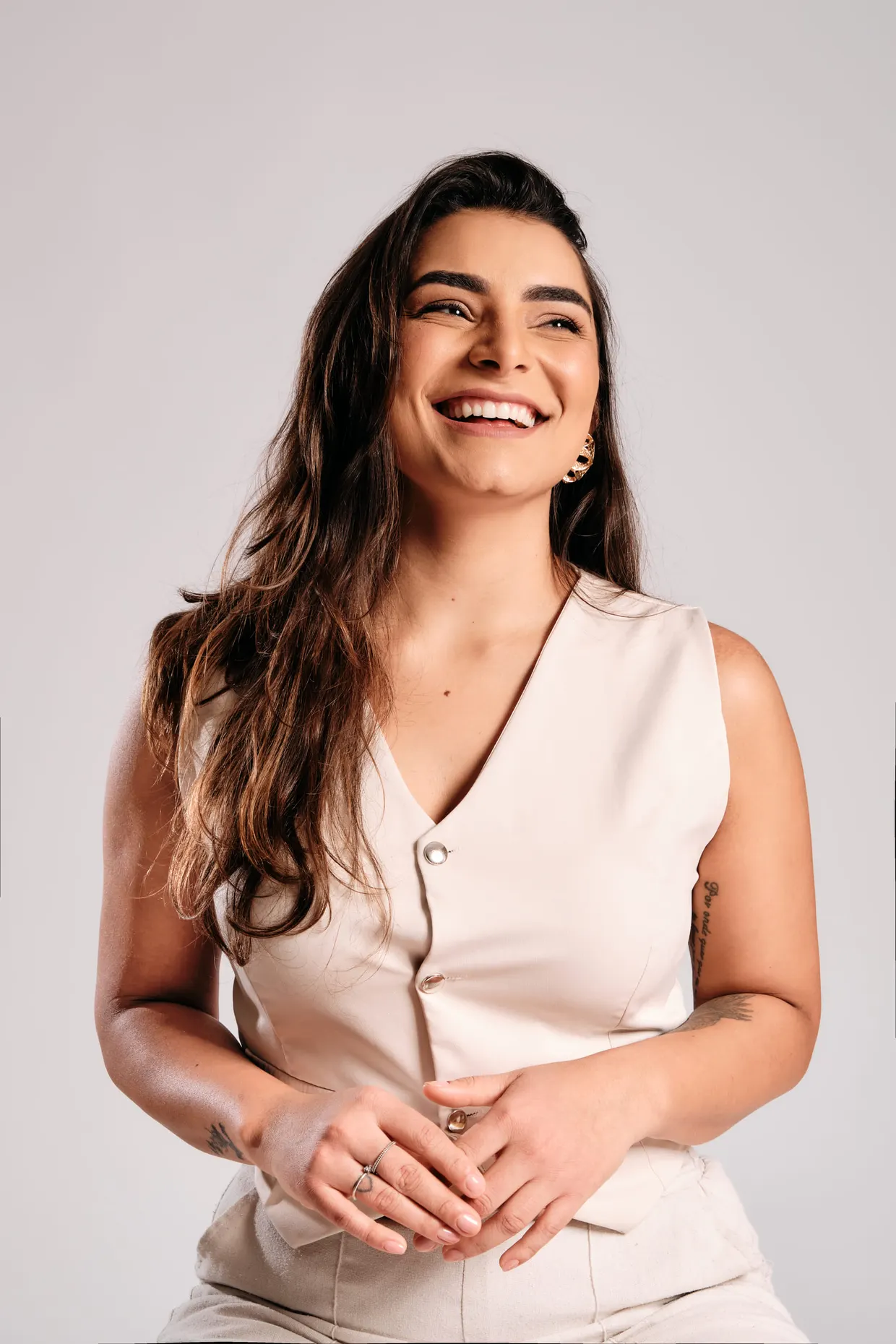 Mulher de colete claro sorri com as mãos unidas diante do corpo.