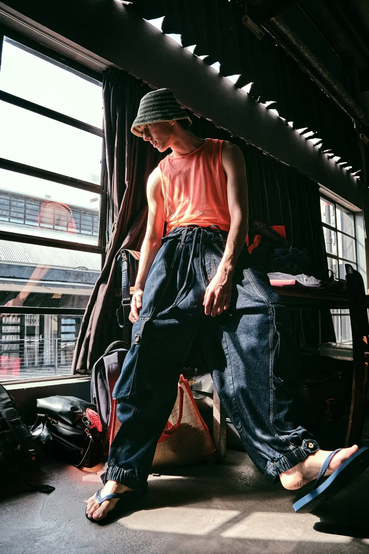 Modelo de chapéu e blusa coral posa junto à janela em um ambiente industrial.
