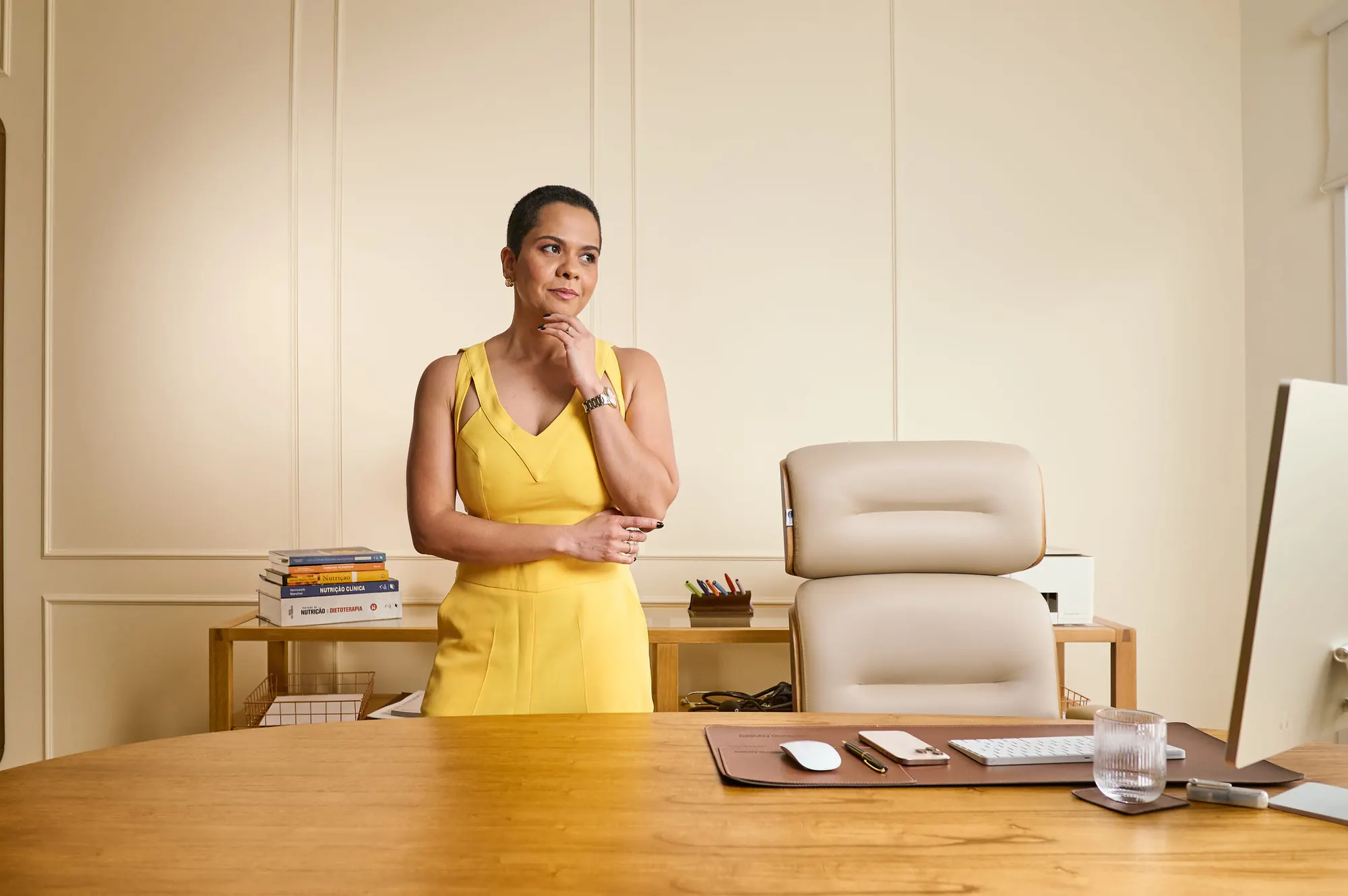 Mulher de vestido amarelo posa atrás de uma mesa em um escritório.