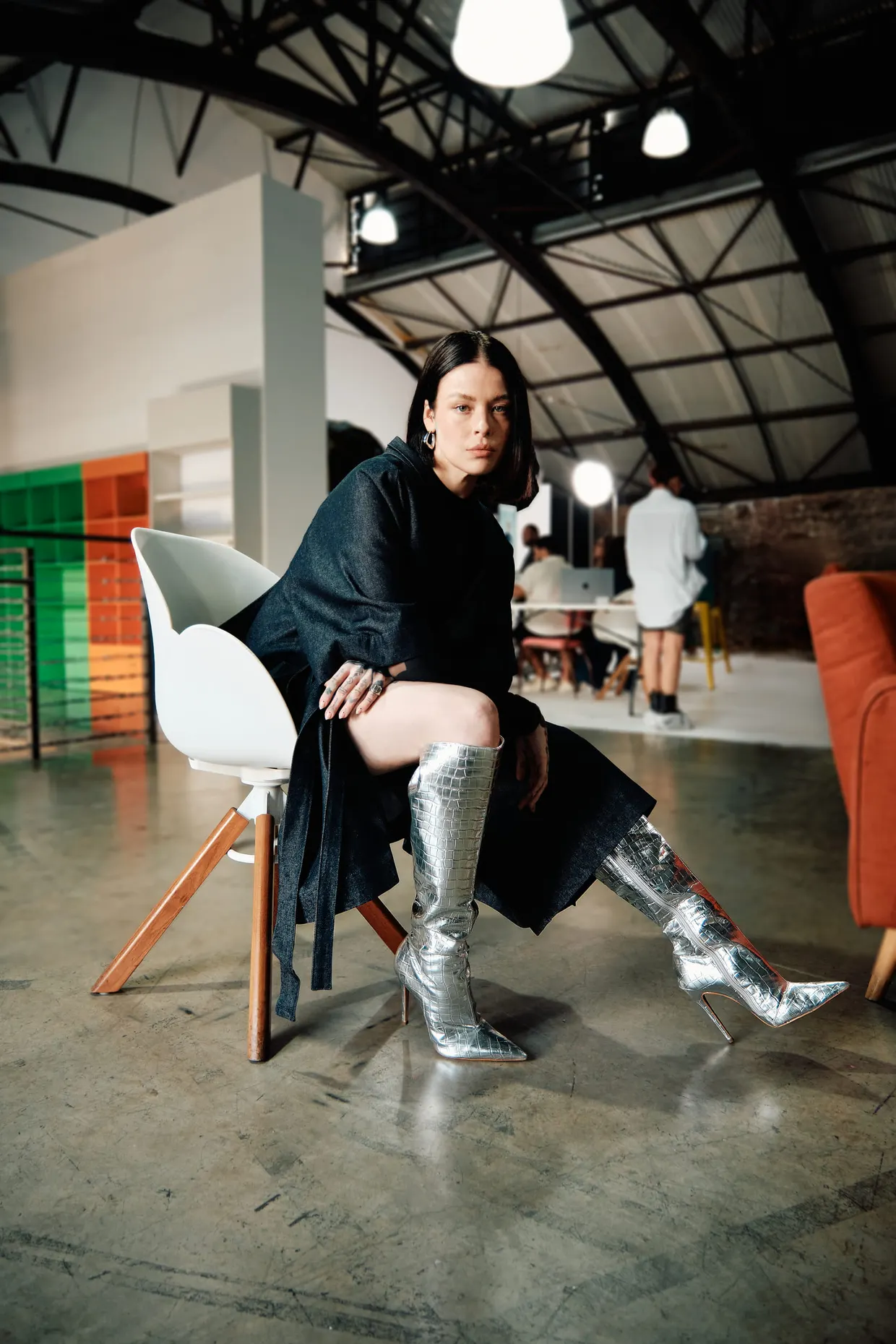 Modelo de casaco escuro e botas prateadas sentado em ambiente industrial.